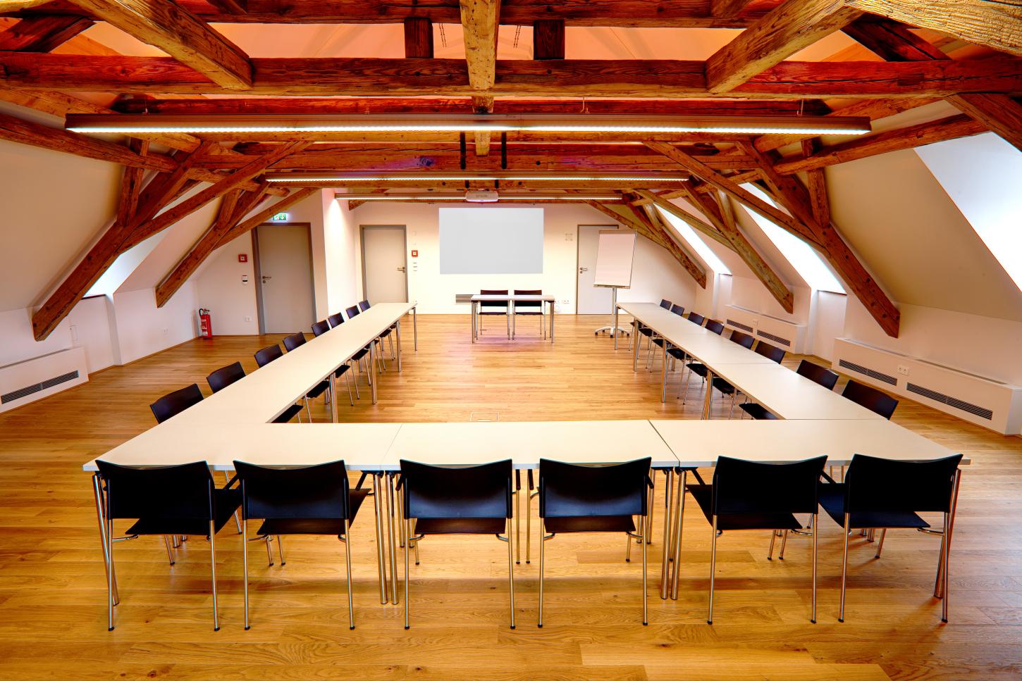 Großer Vortragssaal im Dachgeschoss des Krügerhauses mit offenem Dachstuhl. Im Raum steht eine U-förmige Tafel mit Stühlen vor einer Leinwand mit Beamer.