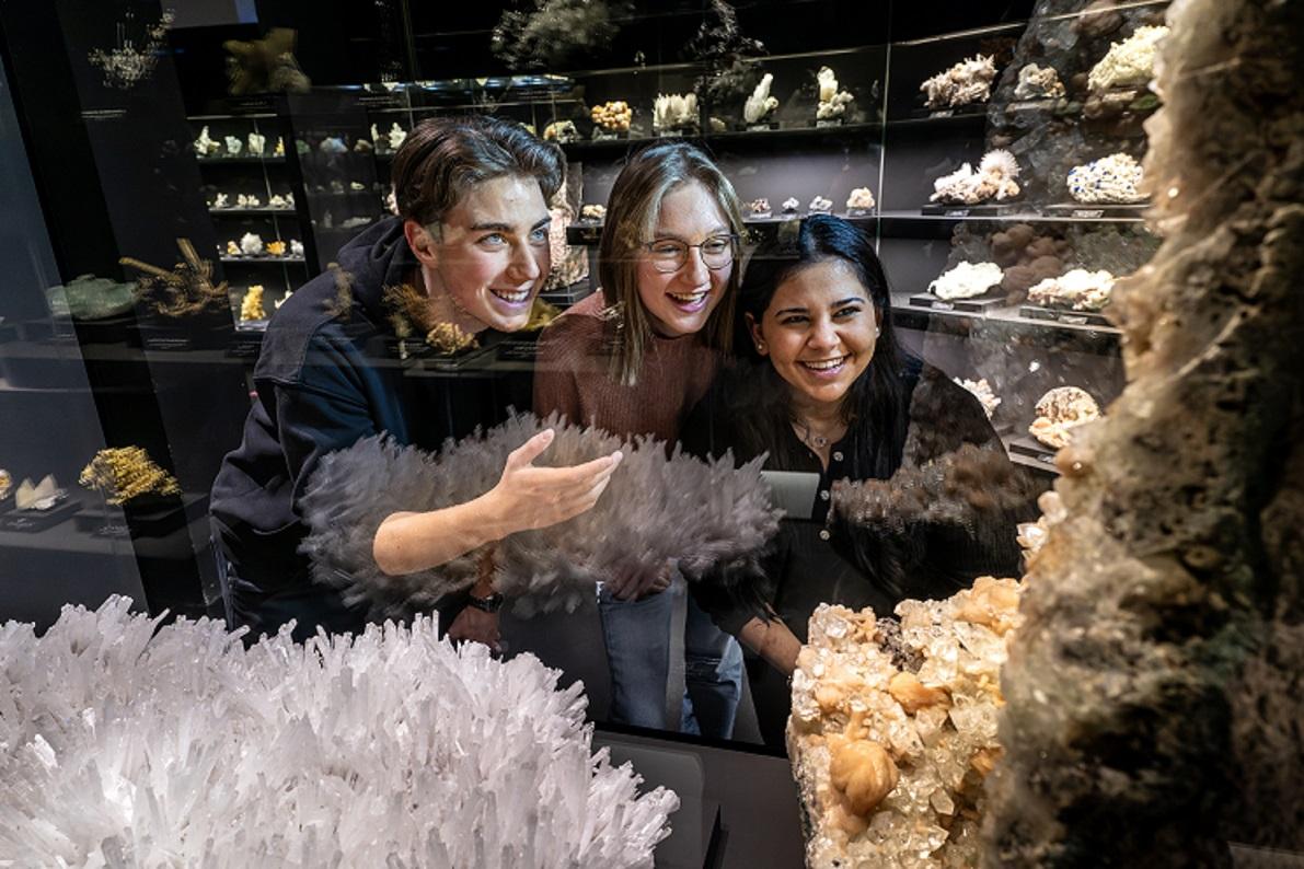 Drei Jugendliche - ein Junge, zwei Mädchen - schauen in der Ausstellung durch eine Vitrine und betrachten zwei große Mineralstufen. Im Hintergrund sieht man weitere kleine Minerale in Vitrinen. 