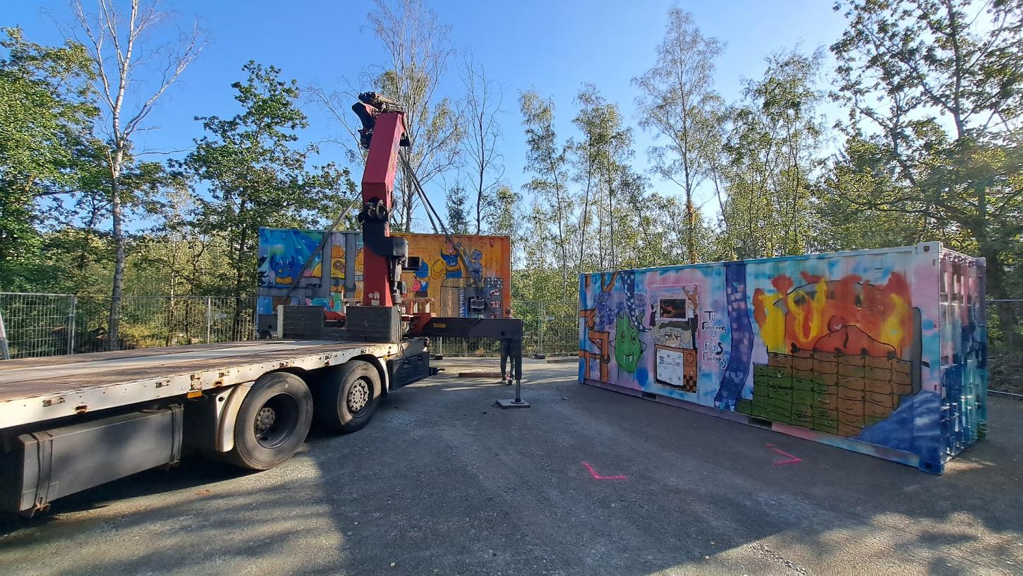 Ein LKW mit Ladekran stellt bunt bemalte Container auf einer begradigten Fläche ab.