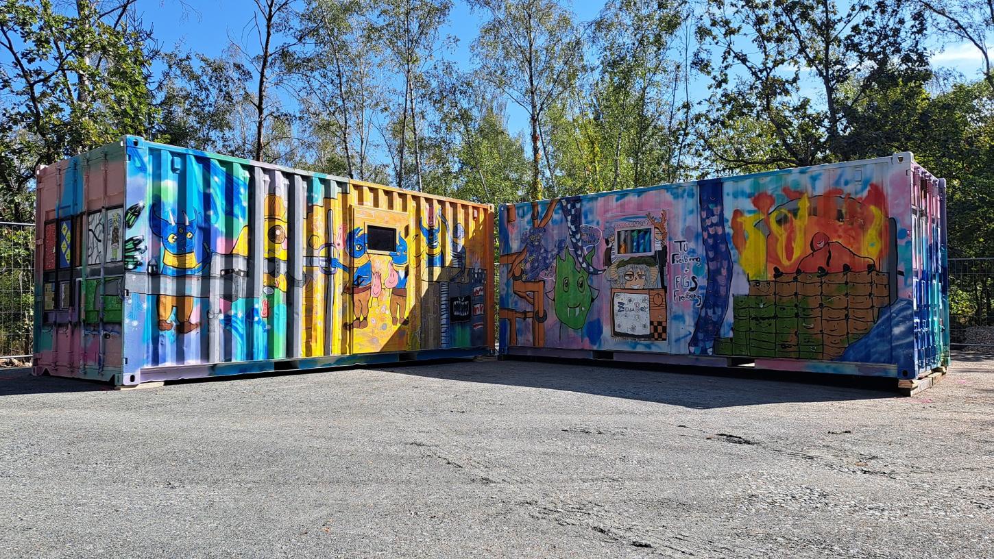 Bunt bemalte Container stehen auf einer begradigten Fläche. Im Hintergrund sind Bäume zu sehen.